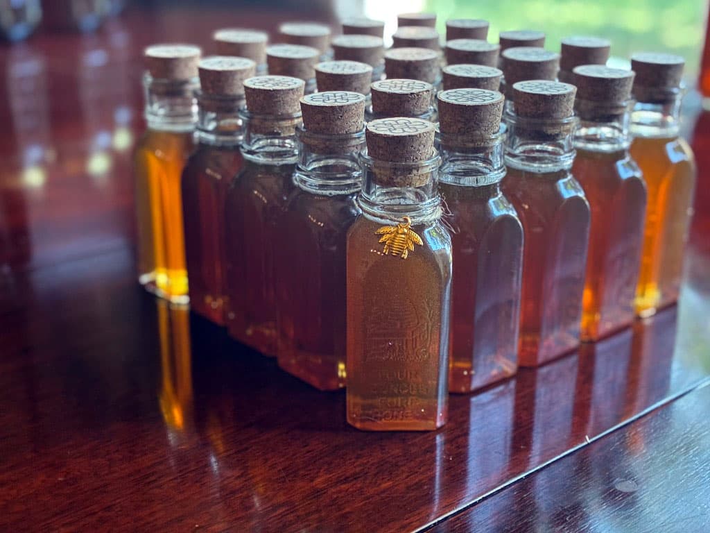 Rows of small square corked honey jars on a wooden table, with a gold bee charm on the jar in front